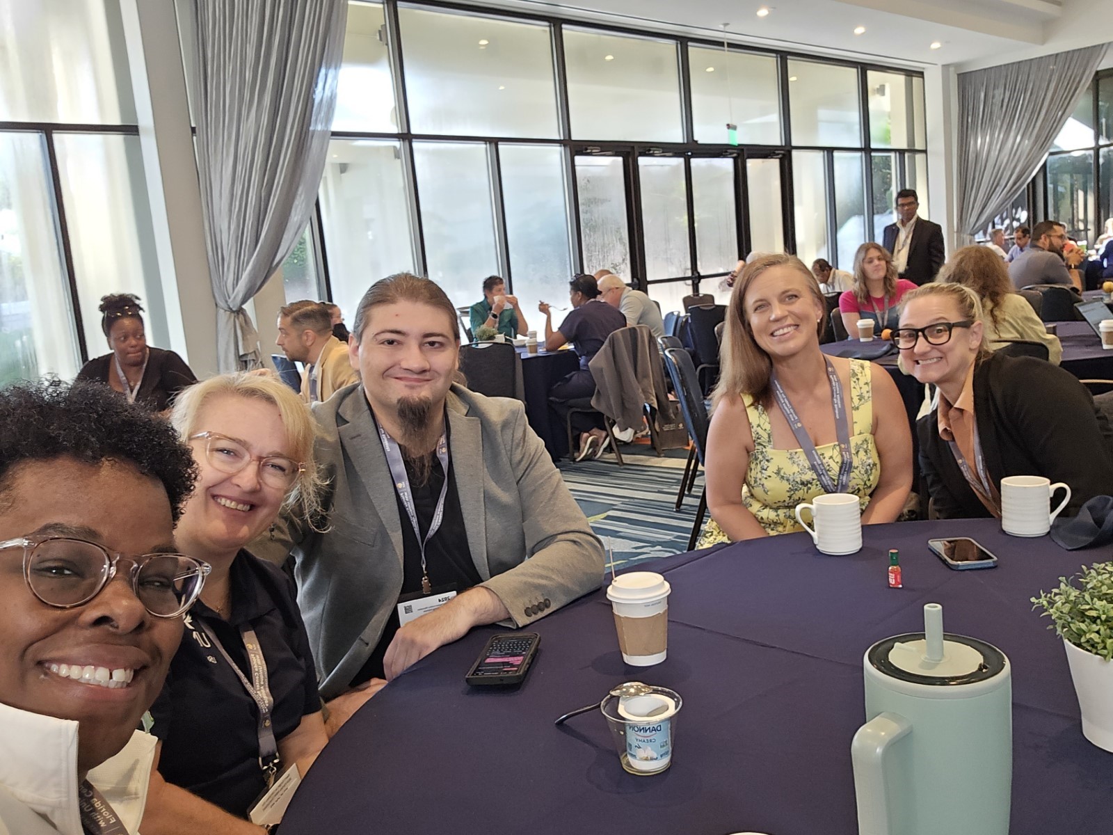 A group of five people are sitting around a round table at a conference or event, smiling for the camera. The setting is a spacious, bright room with large windows and curtains. The group consists of a diverse mix of individuals, with two women on the left, one wearing glasses and the other smiling brightly. A man with a beard and long hair sits in the middle wearing a grey blazer. Two more women are seated to the right, one wearing a yellow floral dress and the other in glasses. The table is set with coffee cups and a small plant, creating a relaxed and collaborative atmosphere. Other attendees are visible in the background, engaged in conversations.